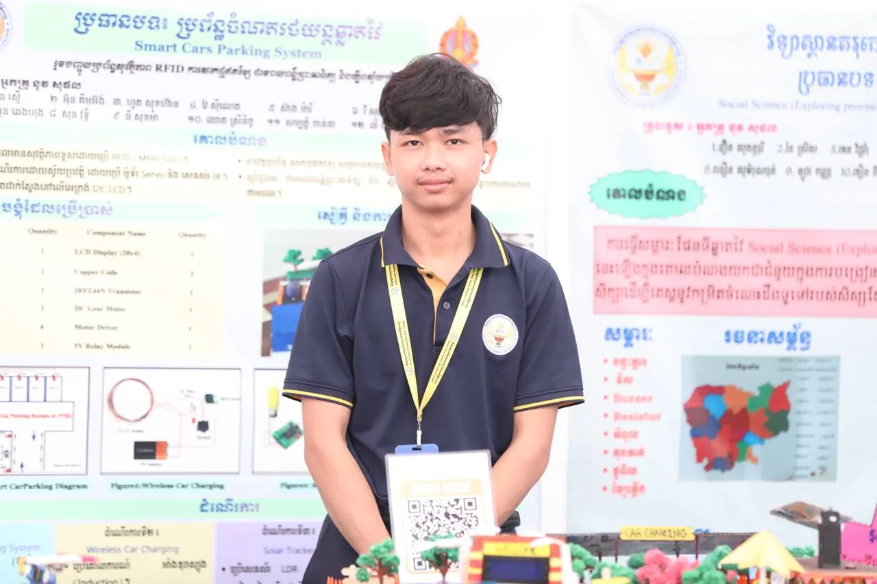 A primary school student teacher wearing a dark blue uniform shirt and a lanyard stands in front of a science fair poster titled "Smart Cars Parking System," holding a scan-able QR code sign.