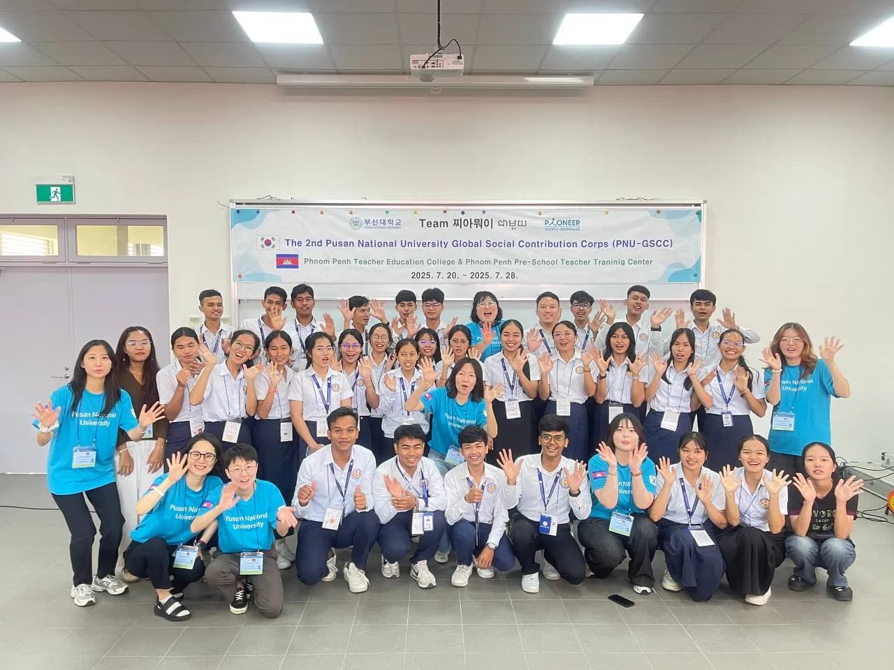A large group photograph of Cambodian student teachers in white and navy uniforms and Korean participants in bright blue t-shirts waving and smiling at the camera. They are gathered indoors in front of a wide banner for the 2nd Pusan National University Global Social Contribution Corps.