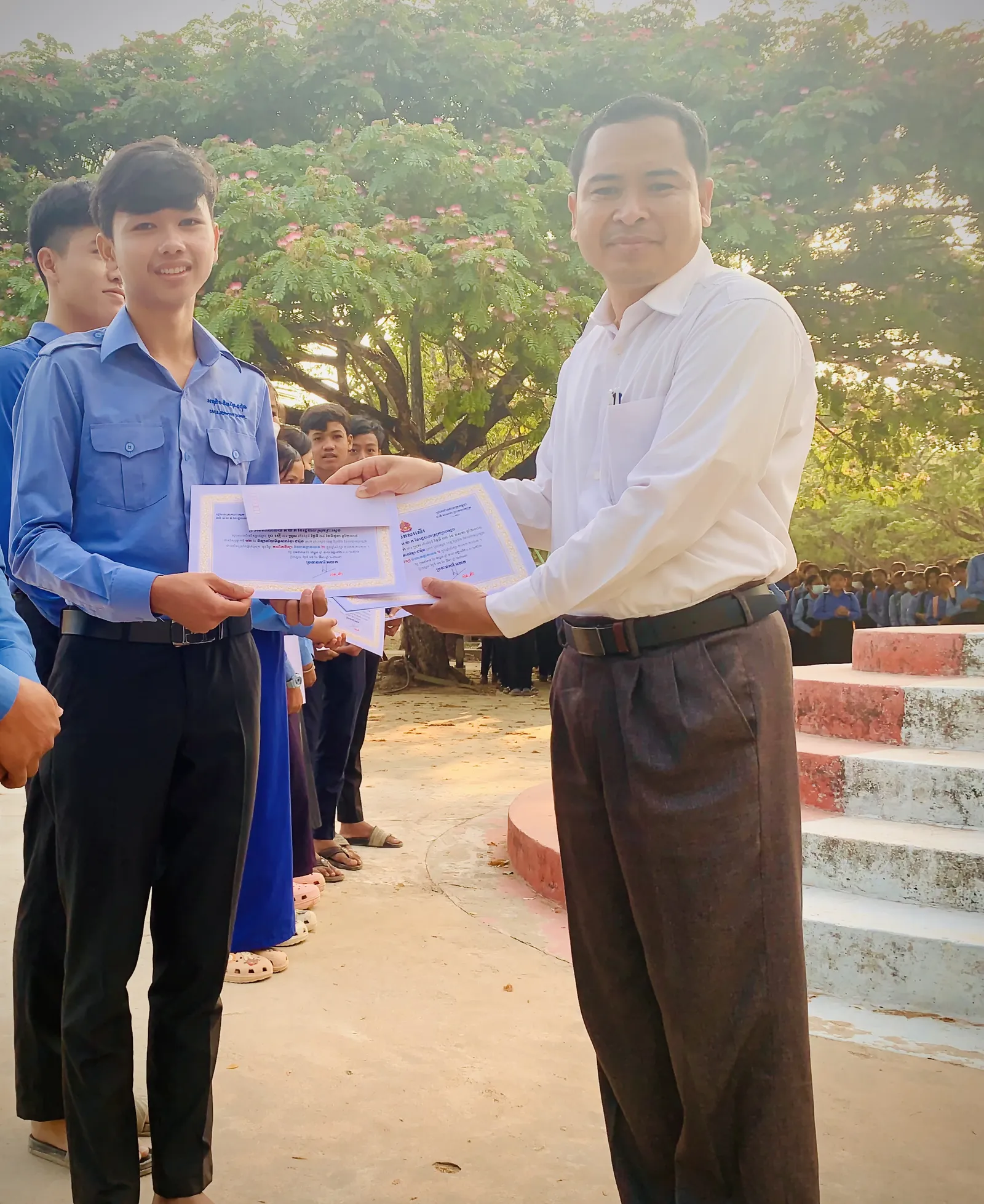 A smiling young man in a blue school uniform receives certificates and a white envelope from a man in a white shirt and brown trousers. They are standing outdoors in a schoolyard with other students and a large tree in the background.