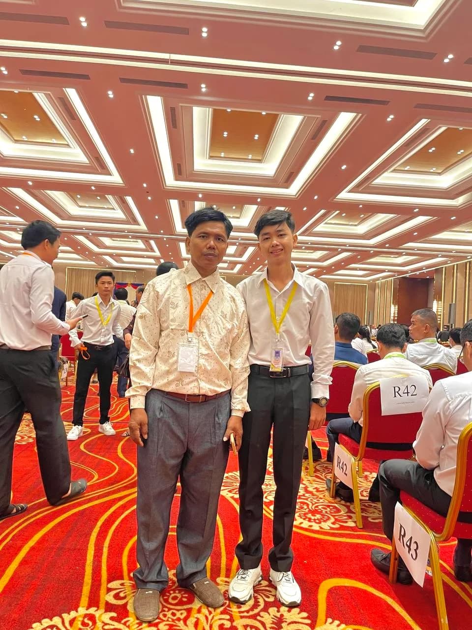 A young man in a white shirt and black trousers stands next to an older man in a patterned shirt inside a large, brightly lit banquet hall with a red carpet.